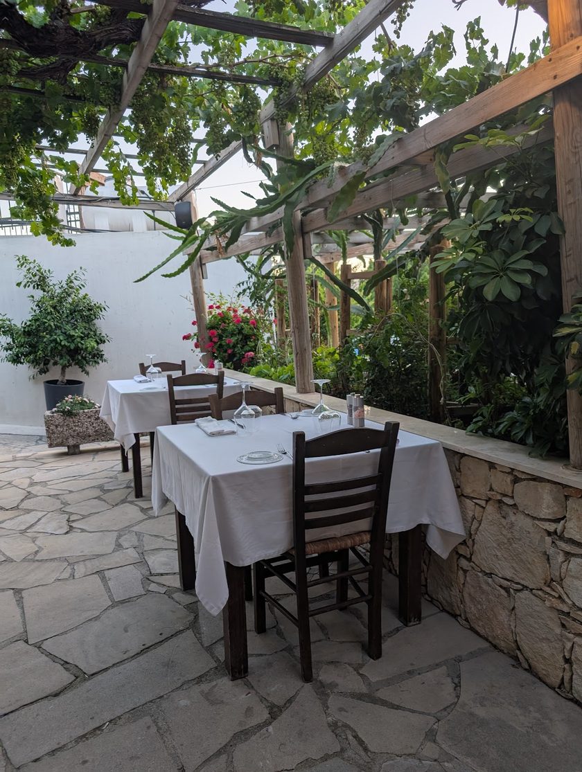 Tables for two in white cloths against a low stone wall, under a timber pergola with vines and a whitewashed wall behind