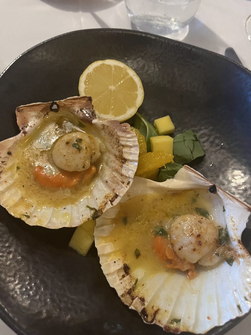 Scallops in their shells in a butter sauce on a dark plate, with diced mango and a lemon wedge
