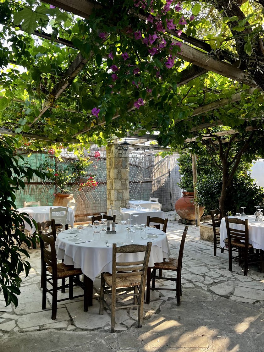 Round tables in white cloths under a vine pergola with pink bougainvillea, rush-seat chairs on stone paving and a clay jar by the far wall