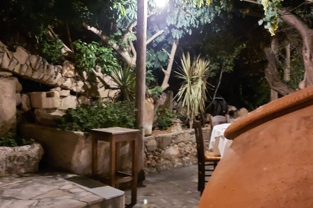 The garden at night under a lamp on a wooden post: stone walls, palms, a small wooden table and a large clay jar in the foreground