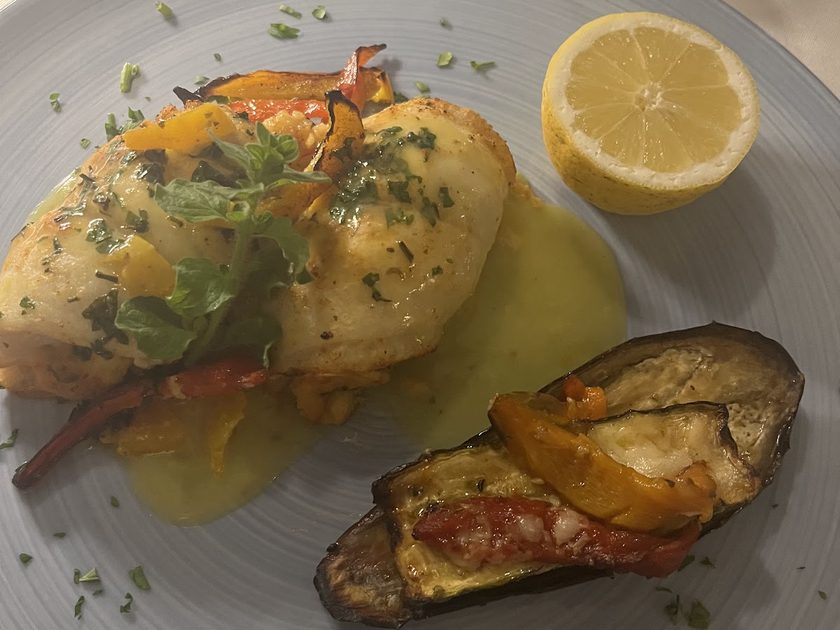 Stuffed sole in a yellow butter sauce with half a lemon and a baked aubergine half, on a grey plate